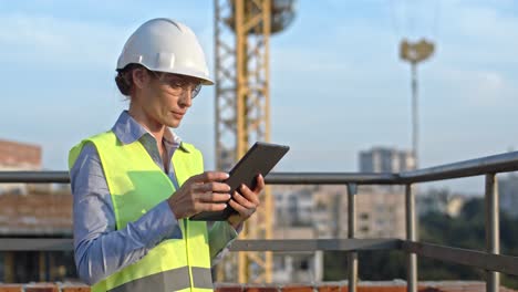 Caucasian-young-beautiful-woman-architect-or-builder-in-the-hardhat-standing-at-the-roof-of-the-building-site-and-using-tablet-device,-tapping-and-scrolling.